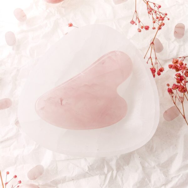 Pembe kuvars gua sha taşı çiçeklerle süslenmiş beyaz arka planda görsellenmiş